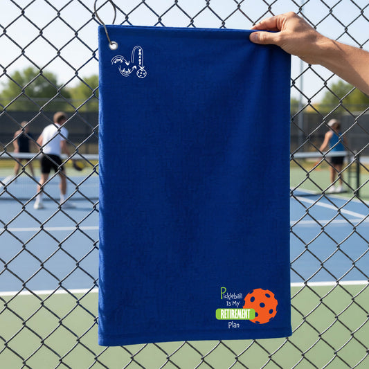 A close-up of a royal blue terry cloth grommet court towel on a pickleball court, with a graphic of the words “Pickleball is my Retirement Plan” in green and white with an orange pickleball illustration in the background.
