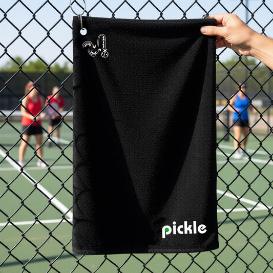 A black textured terry cloth towel with a grommet and hook at the top and a white logo 'DINK DINK SMASH' and a white text 'PICKLE' with a green pickleball inside the letter P, hanging on a fence in front of a pickleball court. 