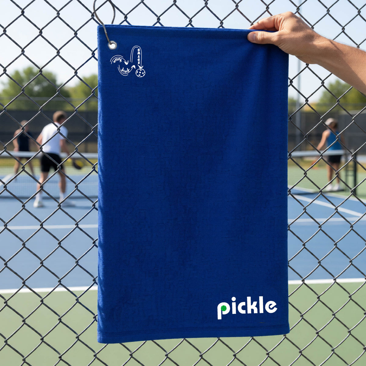 A royal blue textured terry cloth towel with a grommet and hook at the top and a white logo 'DINK DINK SMASH' and a white text 'PICKLE' with a green pickleball inside the letter P, hanging on a fence in front of a pickleball court. 