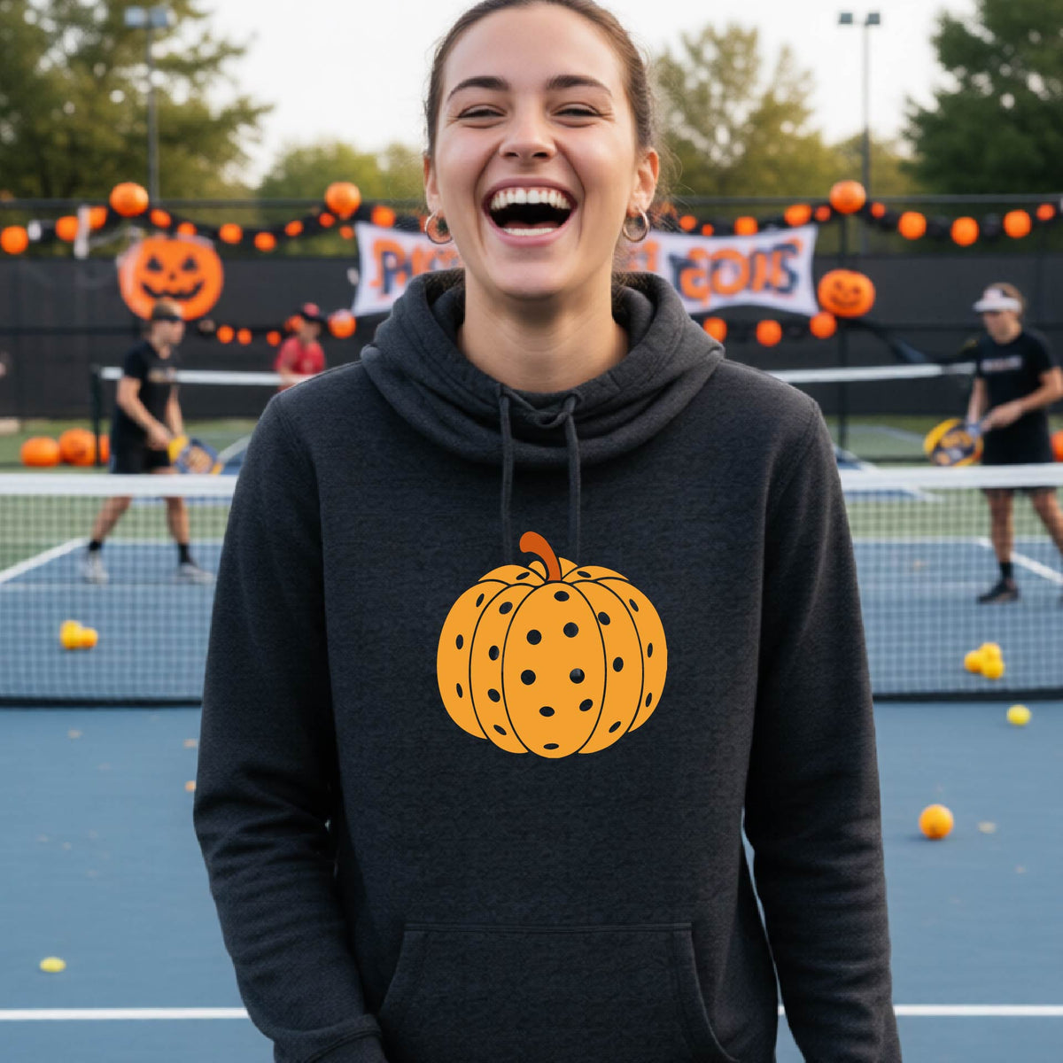 A close-up of a women wearing a black cowl-neck hoodie pullover, with an orange graphic of a pumpkin design like a pickleball,  in front of a pickleball court. 