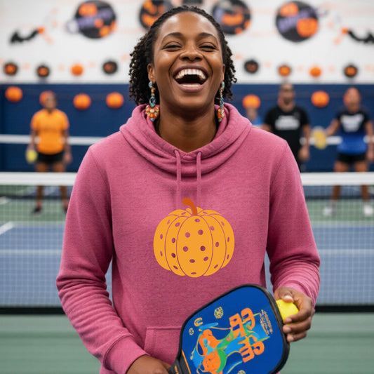 A close-up of a women wearing a pink cowl-neck hoodie pullover, with an orange graphic of a pumpkin design like a pickleball,  in front of a pickleball court. 