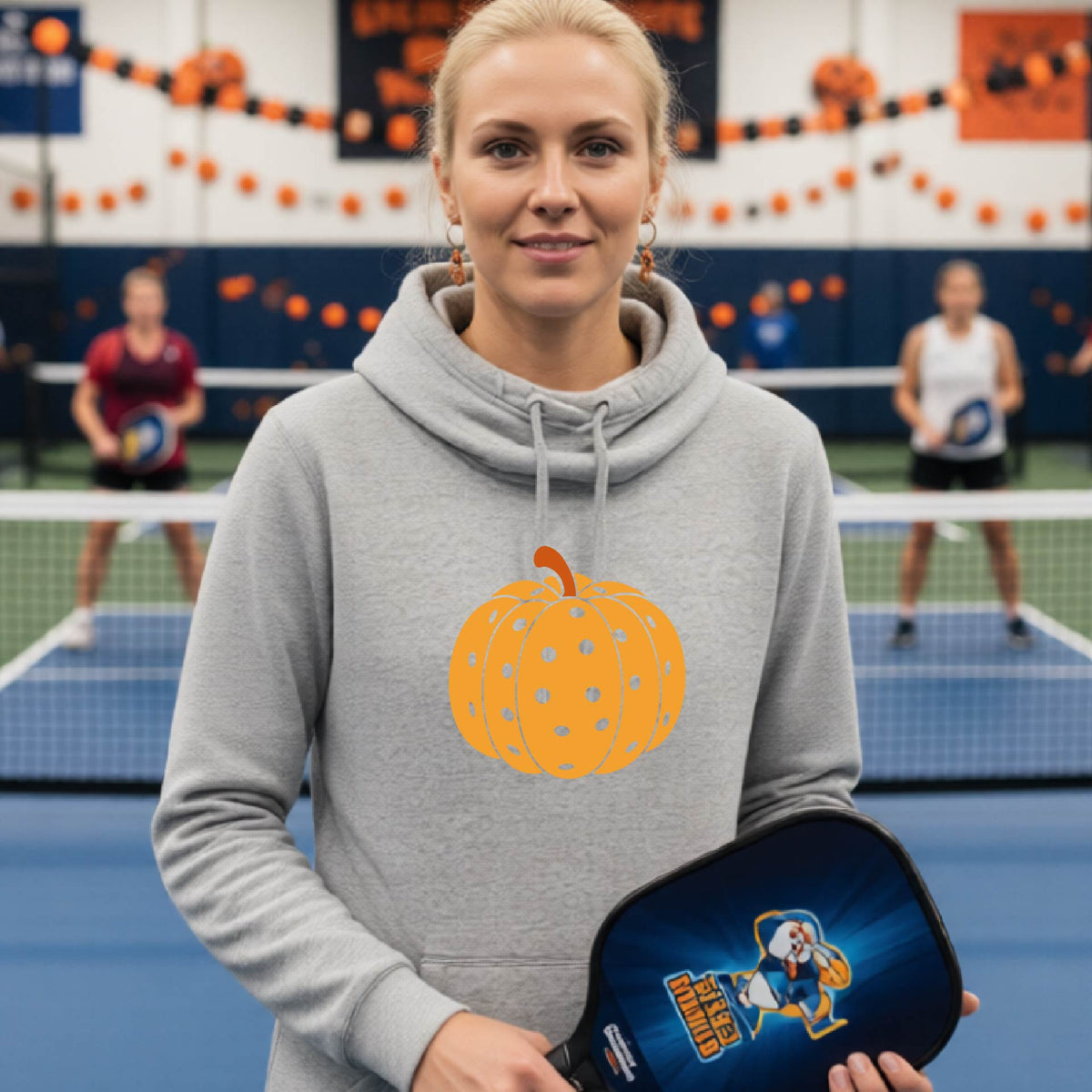 A close-up of a women wearing a gray cowl-neck hoodie pullover, with an orange graphic of a pumpkin design like a pickleball,  in front of a pickleball court. 