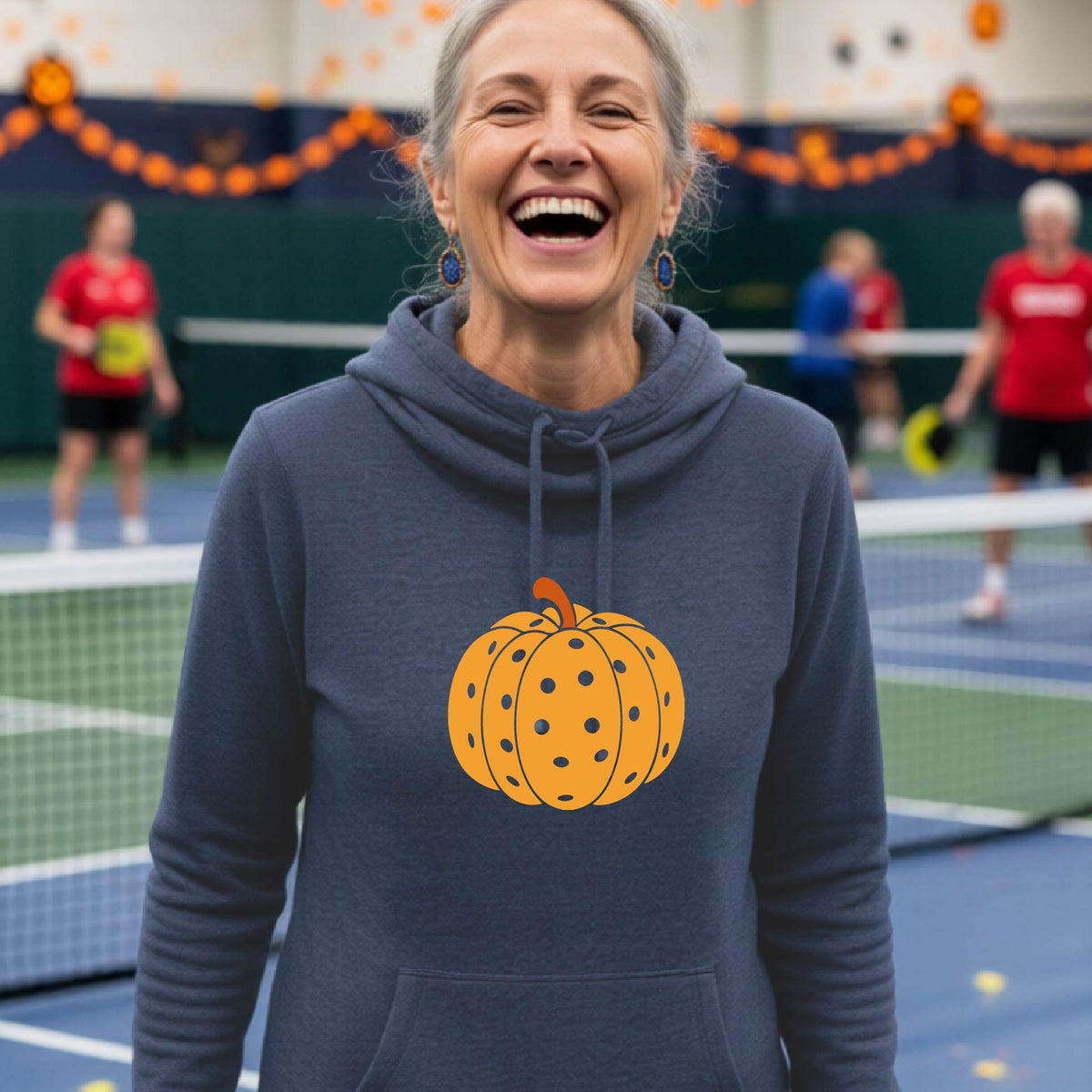 A close-up of a women wearing a navy blue cowl-neck hoodie pullover, with an orange graphic of a pumpkin design like a pickleball,  in front of a pickleball court. 