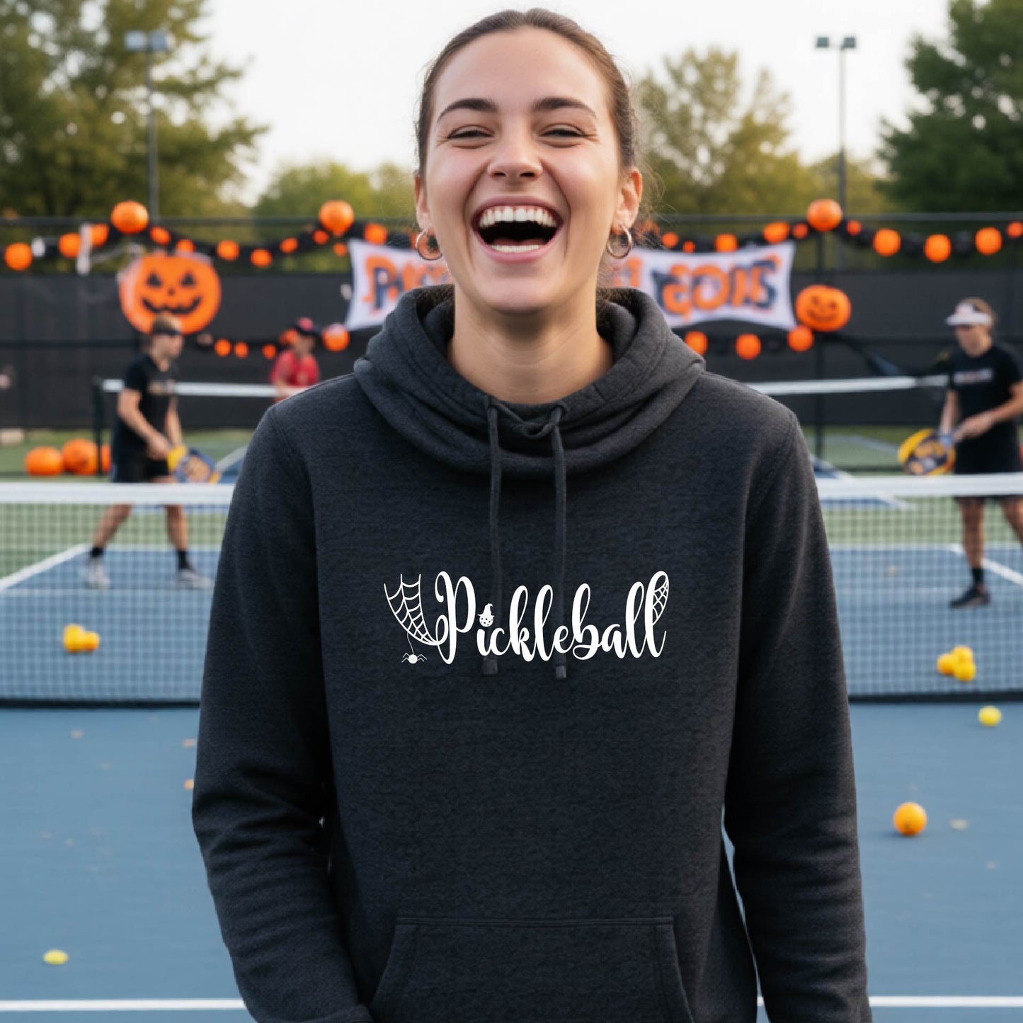 A woman smiling in a dark gray hoodie with a white 'Pickleball' graphic on the chest, standing on an outdoor pickleball court decorated for Halloween.