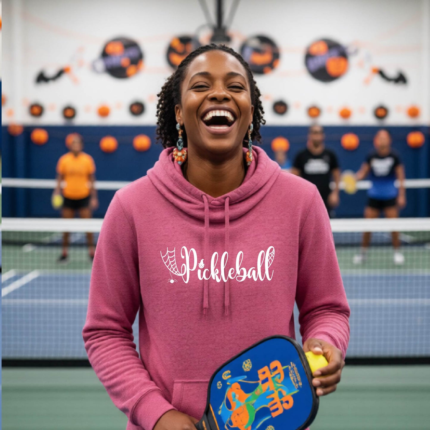 A woman laughing in a pickleball court wearing a pink hoodie with 'Pickleball' printed on the front, holding a blue pickleball paddle and a yellow pickleball.
