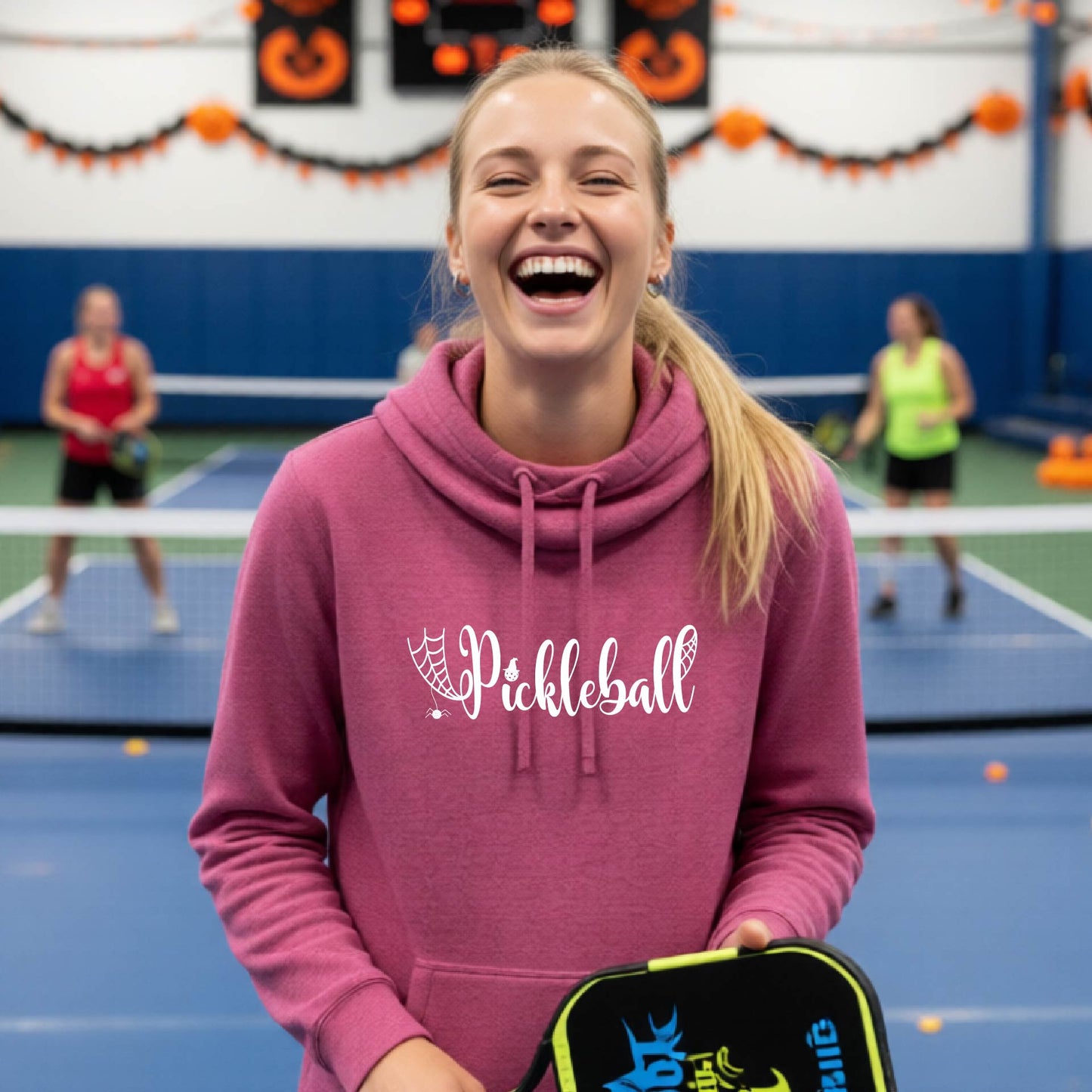 A woman smiling in a pickleball court wearing a pink hoodie with 'Pickleball' printed in white across the chest, holding a pickleball paddle. Other players are visible in the background on a blue court with Halloween decorations.