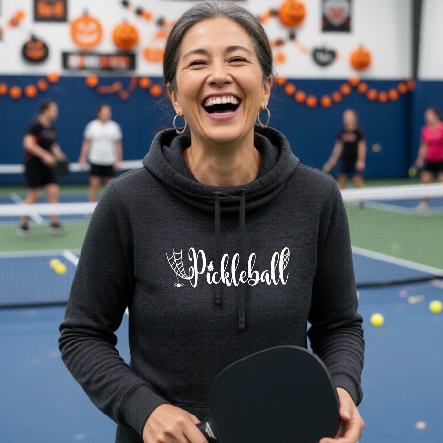 A woman laughing on an indoor pickleball court wearing a black heater hoodie with 'Pickleball' and a spiderweb graphic printed in white on the chest, holding a pickleball paddle.