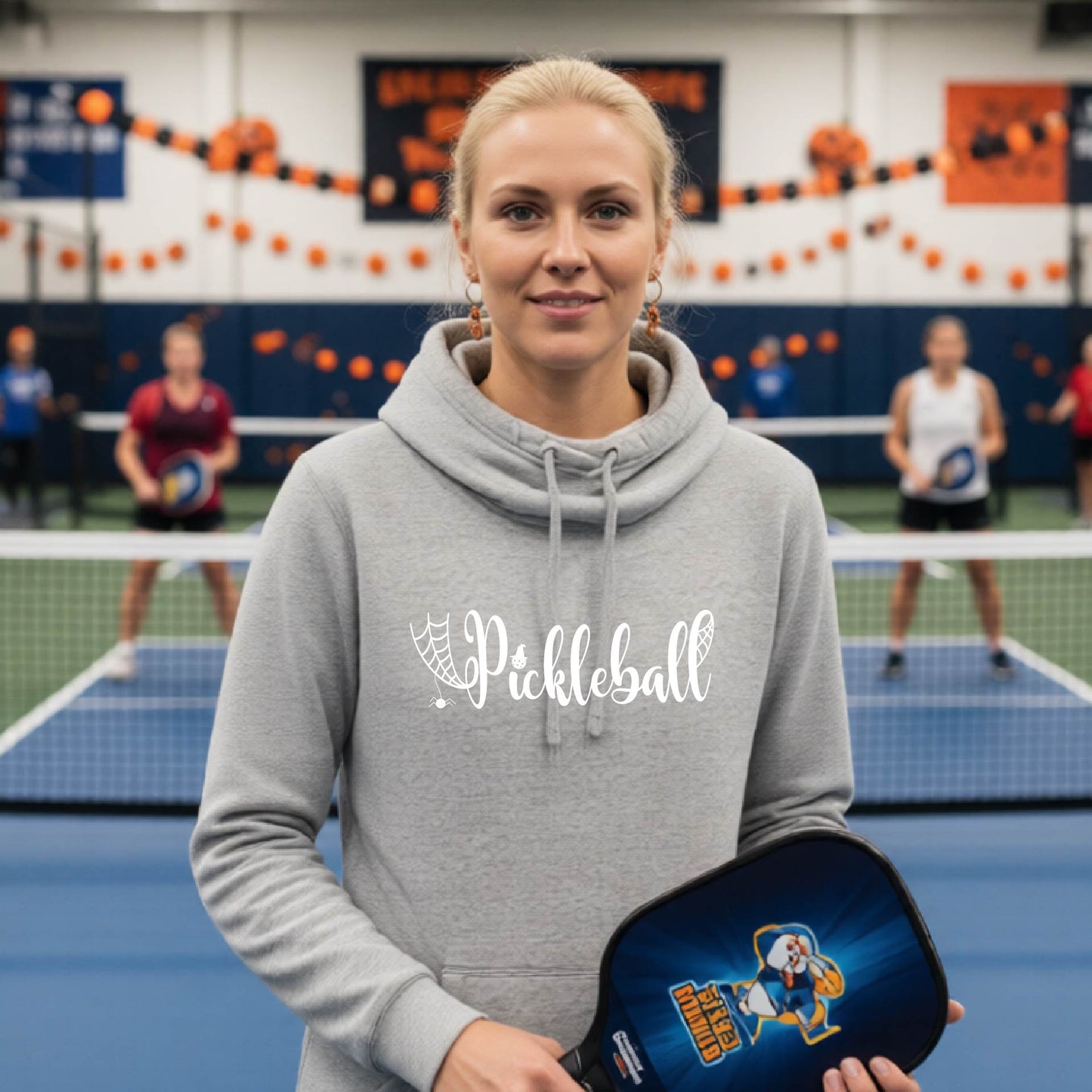 A woman stands on a pickleball court wearing a gray hoodie with 'Pickleball' printed in white across the chest, holding a pickleball paddle. Other players are visible in the background on blue court surfaces with orange and black decorations hanging on the wall.