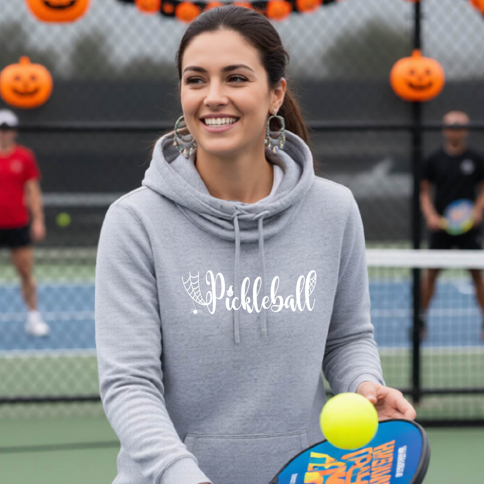 A smiling woman on a pickleball court wearing a gray hoodie with 'Pickleball' printed in white script across the chest, holding a paddle and yellow ball. Pumpkins and a fence are visible in the background.