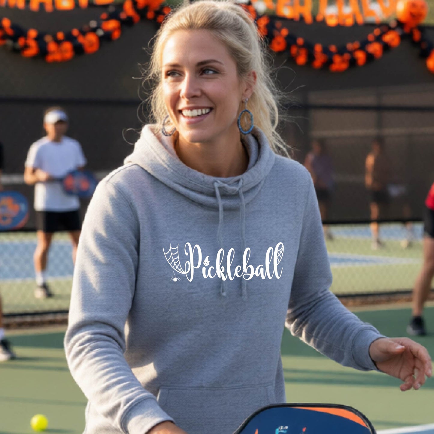 A smiling blonde woman wearing a heather gray hoodie with a white 'Pickleball' graphic on the chest, standing on an outdoor pickleball court with other players and orange/black decorations in the background.