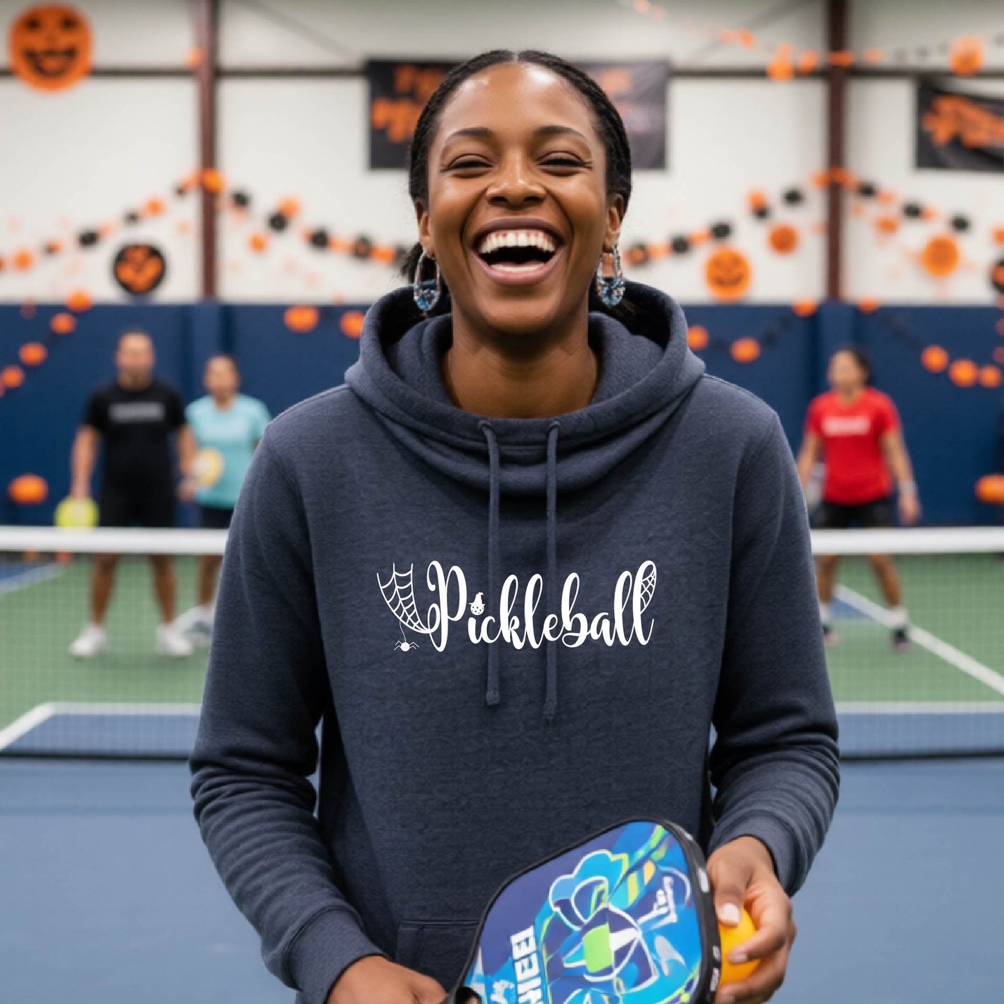 A woman smiling in a gym while wearing a navy blue hoodie with 'Pickleball' printed in white script on the front. She is holding a pickleball paddle in a court setting with other players and orange/black Halloween decorations in the background.