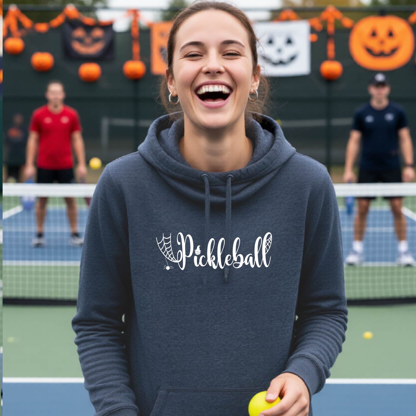 A woman smiling on a pickleball court wearing a navy blue hoodie with a white 'Pickleball' graphic on the chest, holding a yellow pickleball.