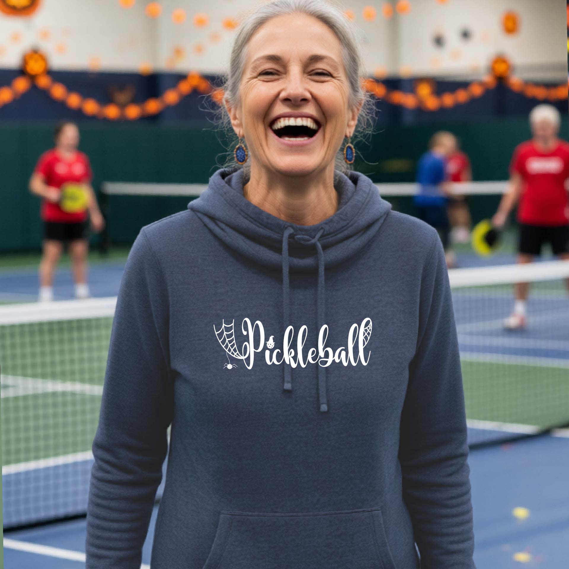A smiling woman wearing a navy blue hoodie with a white 'Pickleball' graphic on the chest, standing on an indoor pickleball court with other players blurred in the background.
