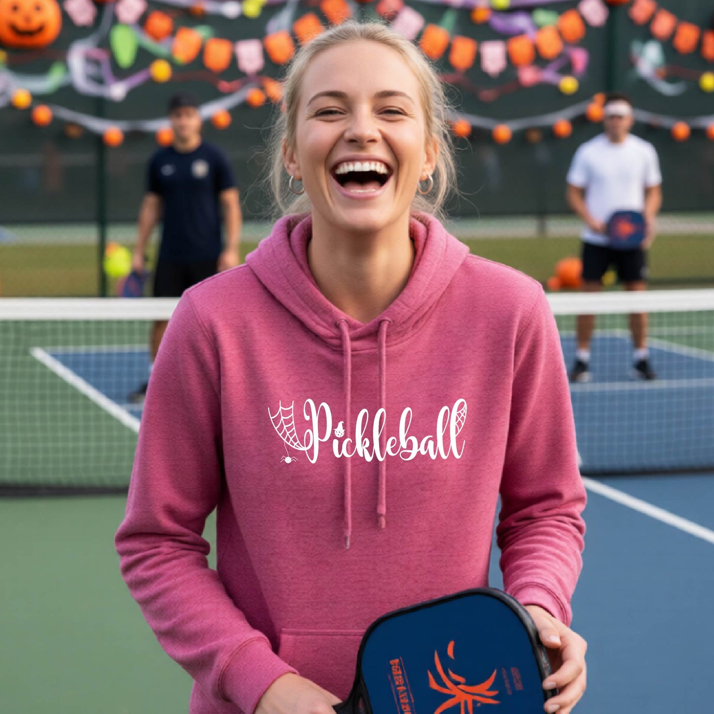 A woman laughing on an outdoor pickleball court wears a pink hoodie with 'Pickleball' printed in white across the chest, holding a blue pickleball paddle case. Other players and festive court decorations are blurred in the background.