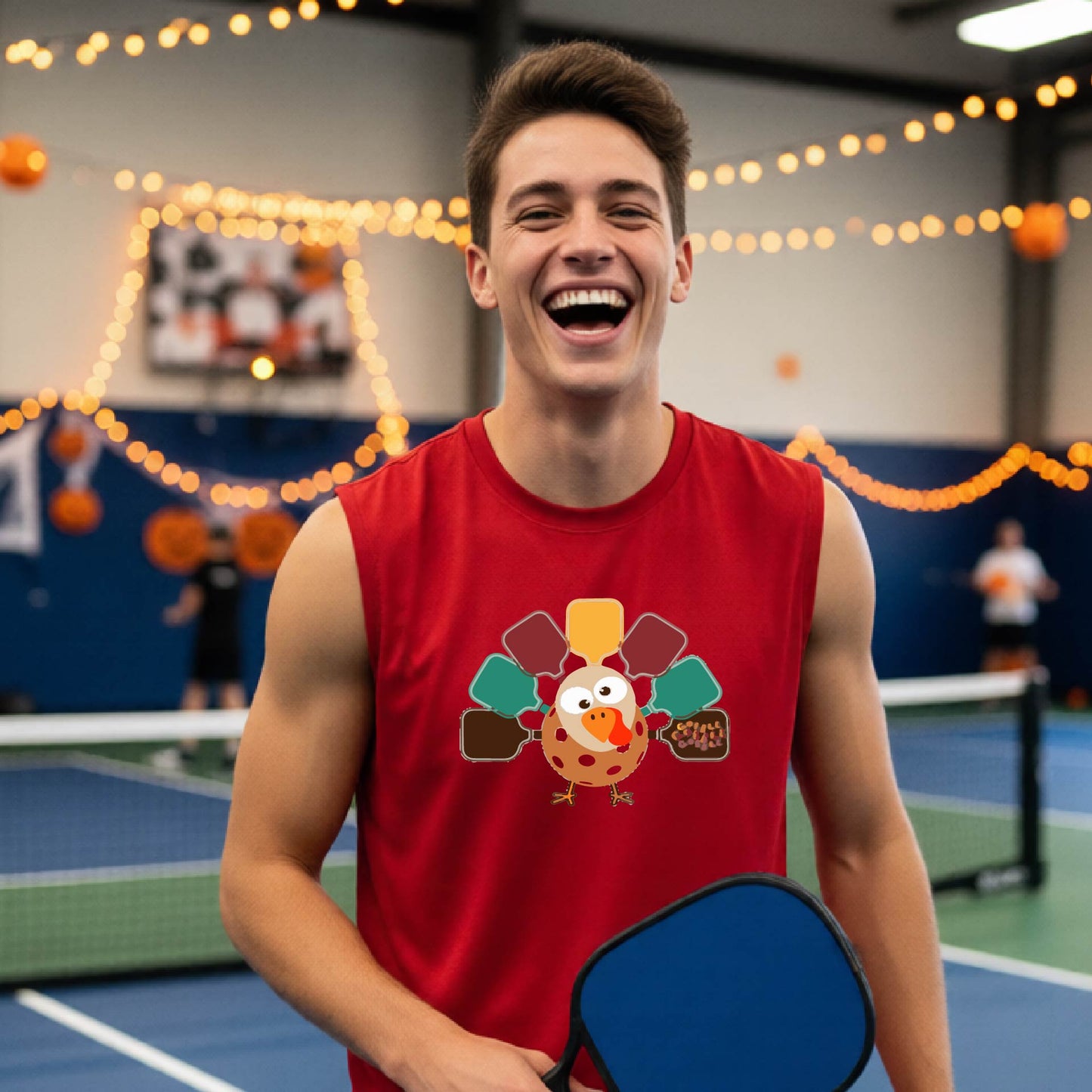 A man standing on a pickleball court smiling and wearing a red sleeveless t-shirt with a pickleball turkey graphic made of pickleblal paddles. 