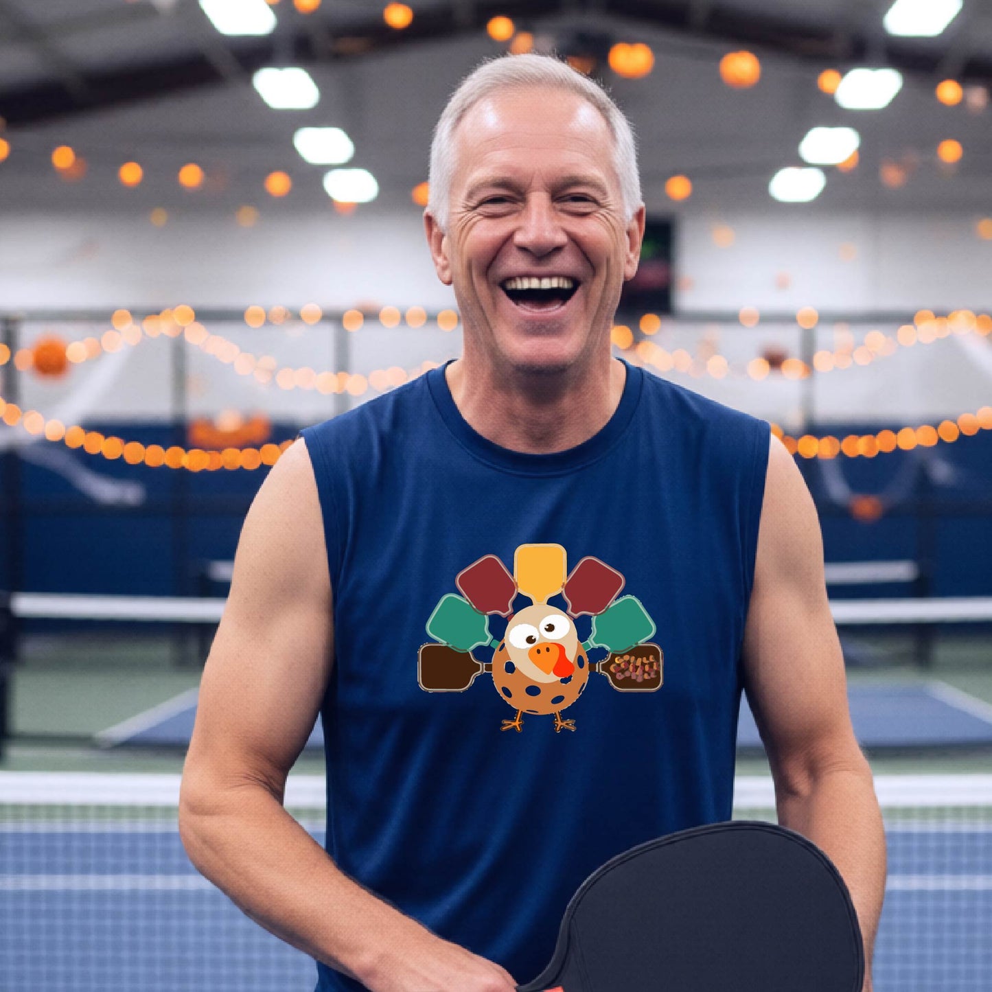 A man standing on a pickleball court smiling and wearing a royal  blue sleeveless t-shirt with a pickleball turkey graphic made of pickleblal paddles. 