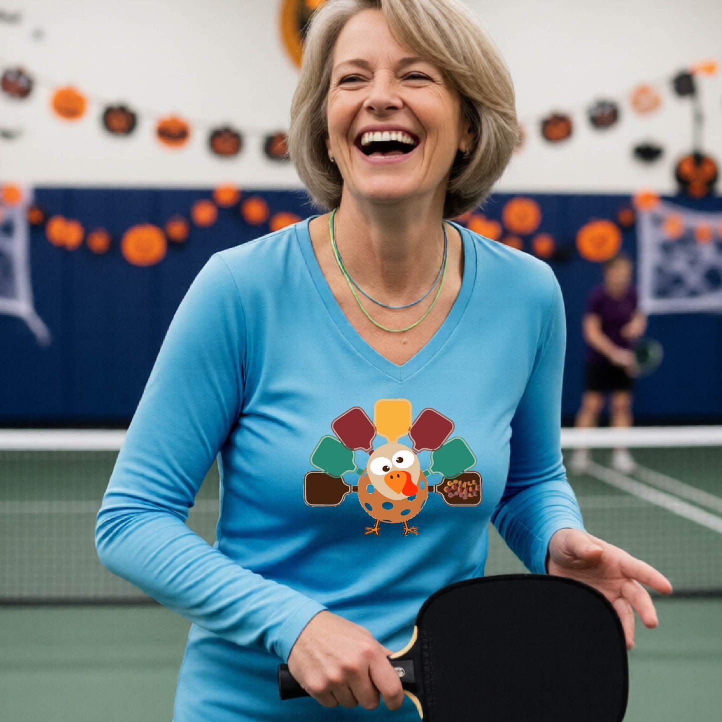 A woman laughing on an indoor paddle ball court wearing a bright blue long-sleeve shirt with a colorful cartoon pickleball turkey graphic on the chest.