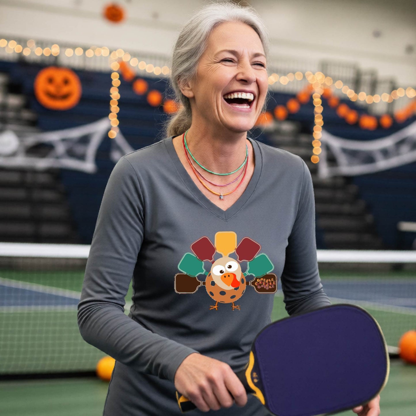 A woman laughing in an indoor sports hall while wearing a gray long-sleeve shirt with a colorful Thanksgiving pickleball turkey graphic on the front, with Halloween decorations like pumpkins and string lights visible in the background.