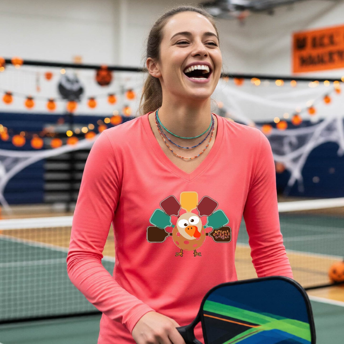 A woman laughing on an indoor pickleball court wearing a coral pink long-sleeve V-neck shirt with a colorful cartoon turkey graphic on the chest.