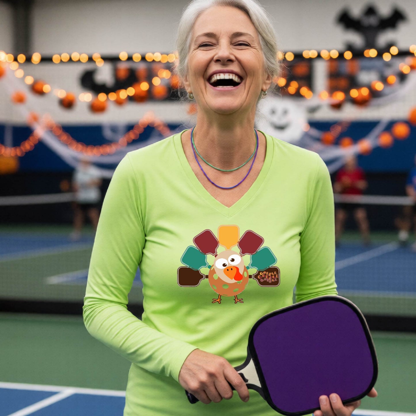 A woman laughing on an indoor pickleball court wearing a lime green long-sleeve V-neck shirt with a colorful cartoon turkey graphic on the chest.