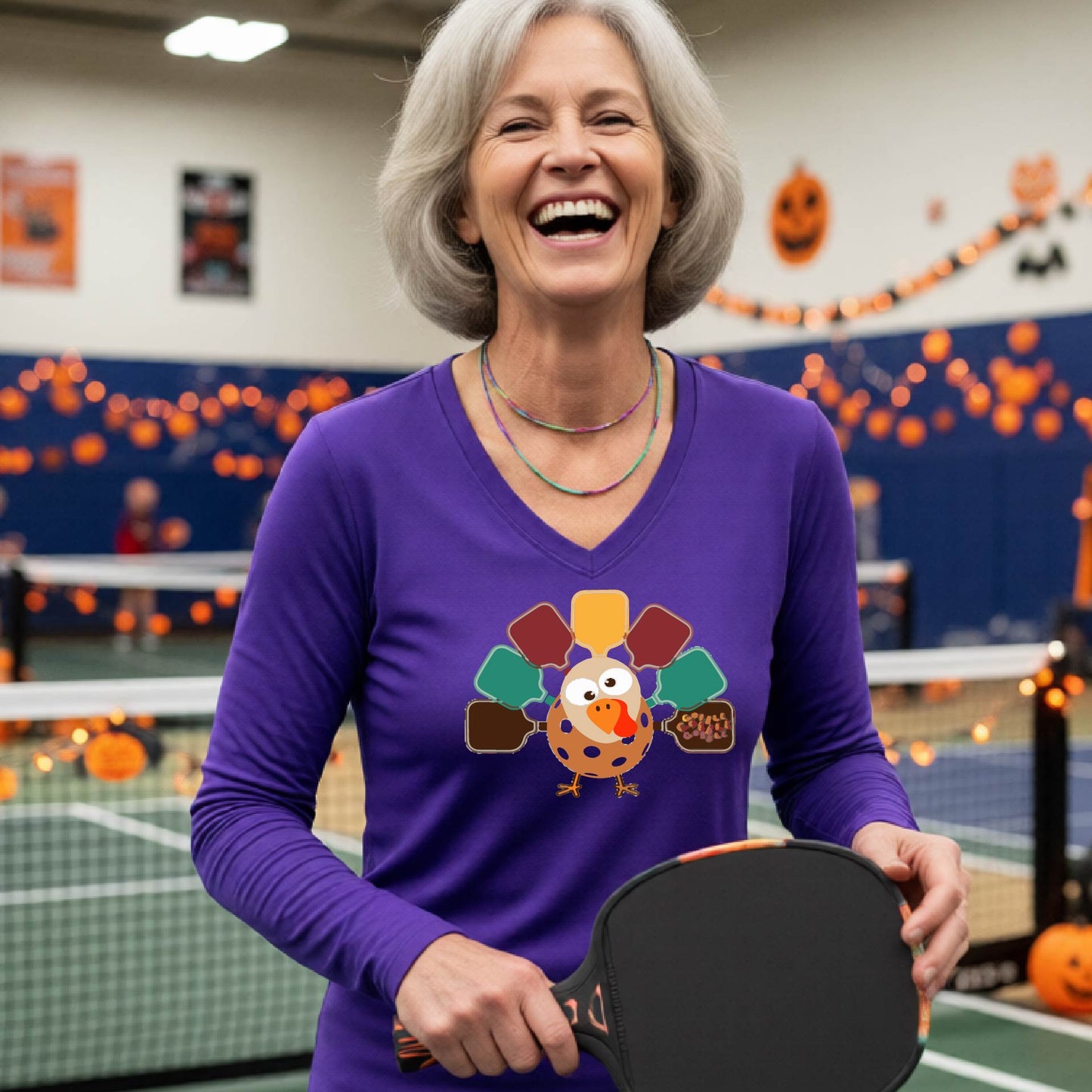 A woman laughing on an indoor pickleball court wearing a purple long-sleeve V-neck shirt with a colorful cartoon turkey graphic on the chest.