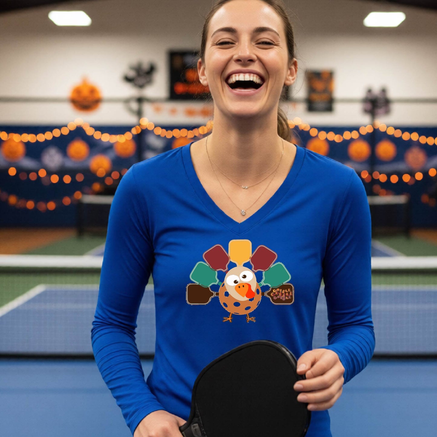 A woman laughing in a pickleball court wearing a royal blue long-sleeve V-neck shirt with a colorful cartoon pickleball turkey graphic on the chest, holding a paddle.