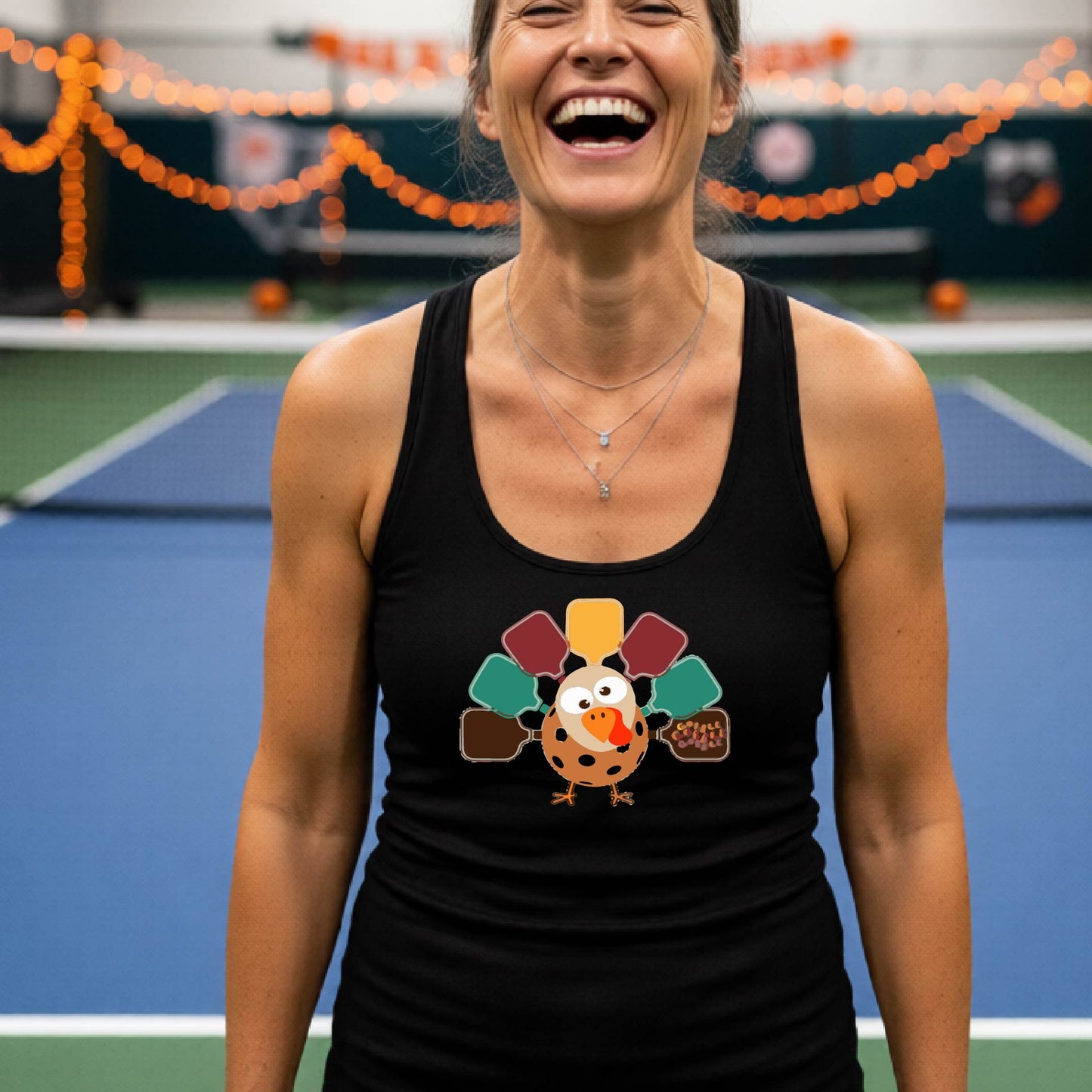 A black racerback tank top featuring a graphic of a cartoon turkey. The turkey's tail feathers are depicted as colorful pickleball paddles in shades of yellow, maroon, teal, and brown,.