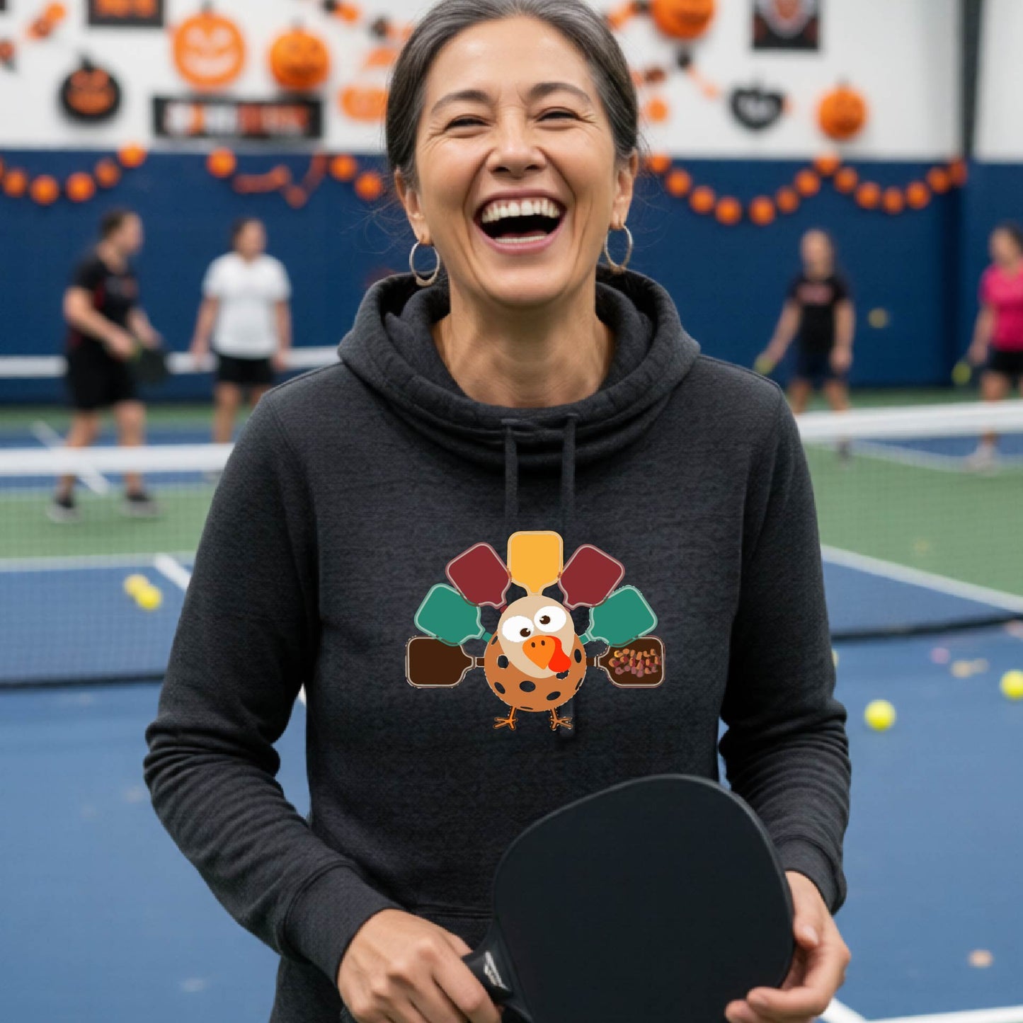 A woman smiling in a pickleball court wearing a black hoodie with a colorful pickleball turkey graphic made of pickleball paddles on the 