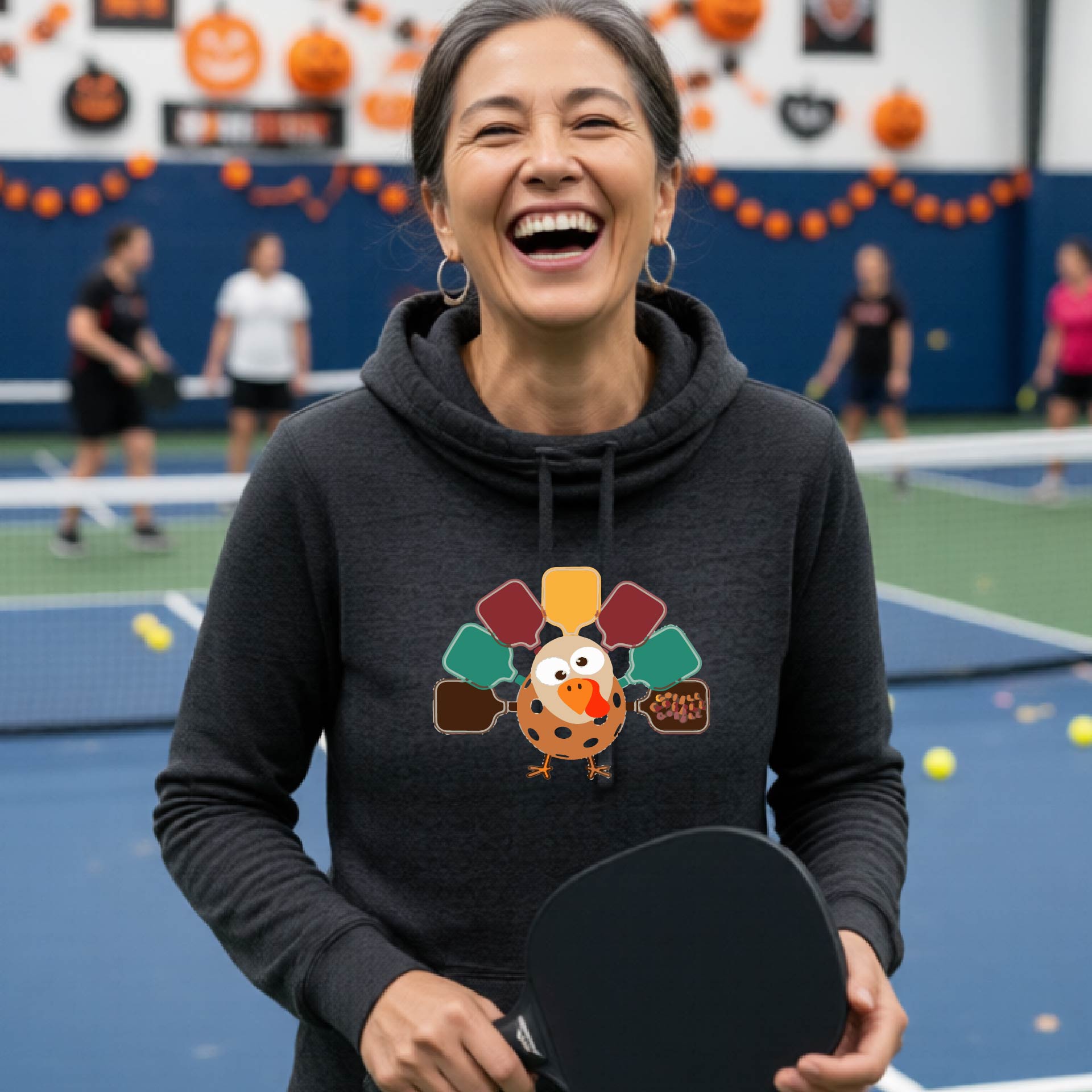 A woman smiling in a pickleball court wearing a black hoodie with a colorful pickleball turkey graphic made of pickleball paddles on the 
