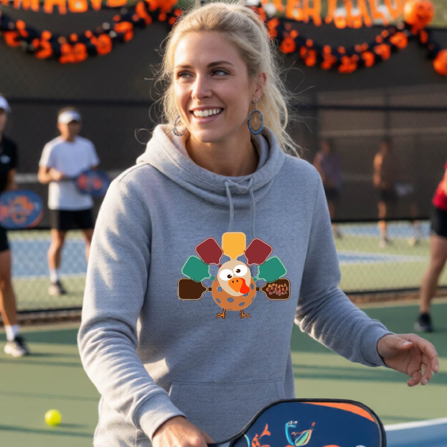 A woman smiling in a pickleball court wearing a gray hoodie with a colorful pickleball turkey graphic made of pickleball paddles on the 