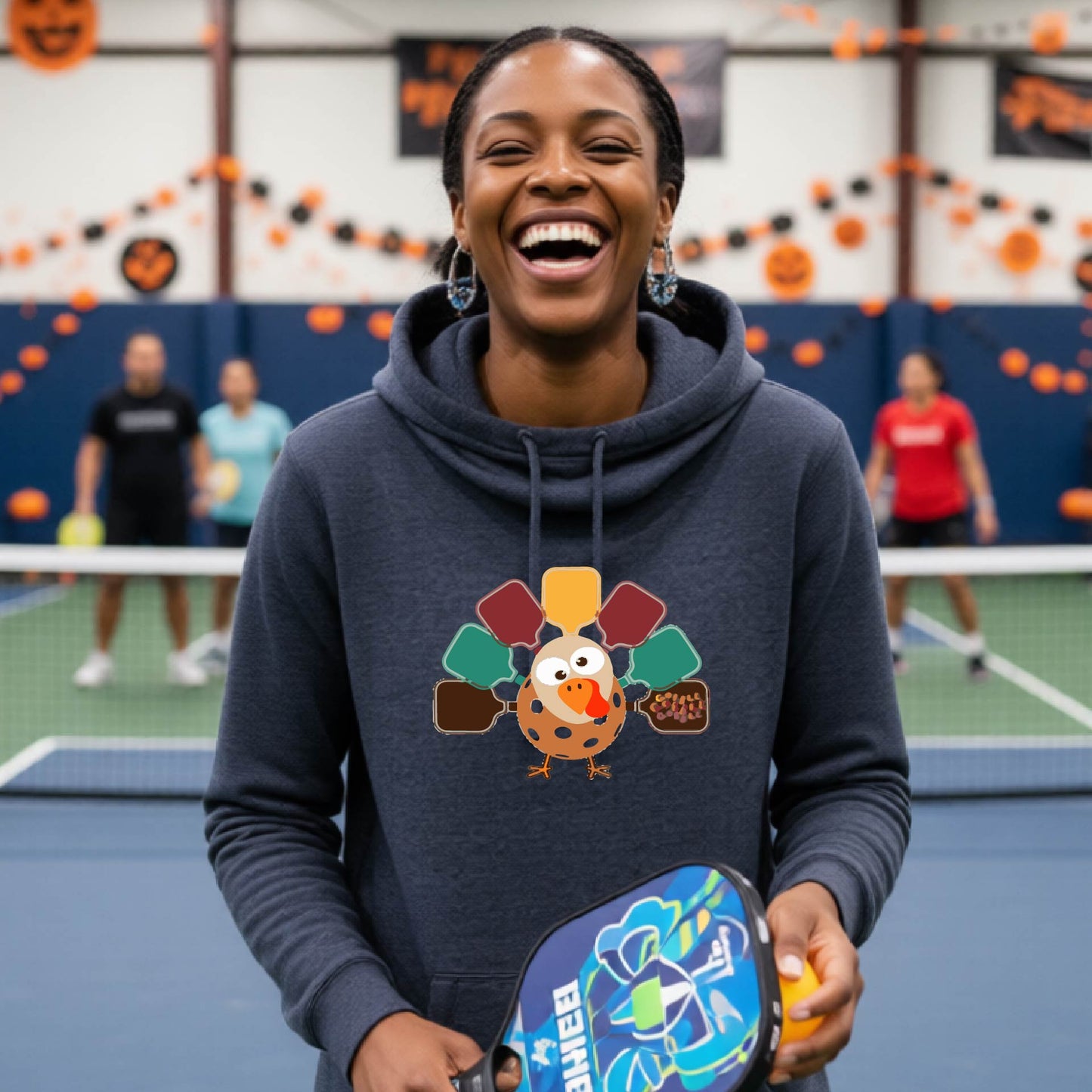 A woman smiling in a pickleball court wearing a navy blue hoodie with a colorful pickleball turkey graphic made of pickleball paddles on the front.