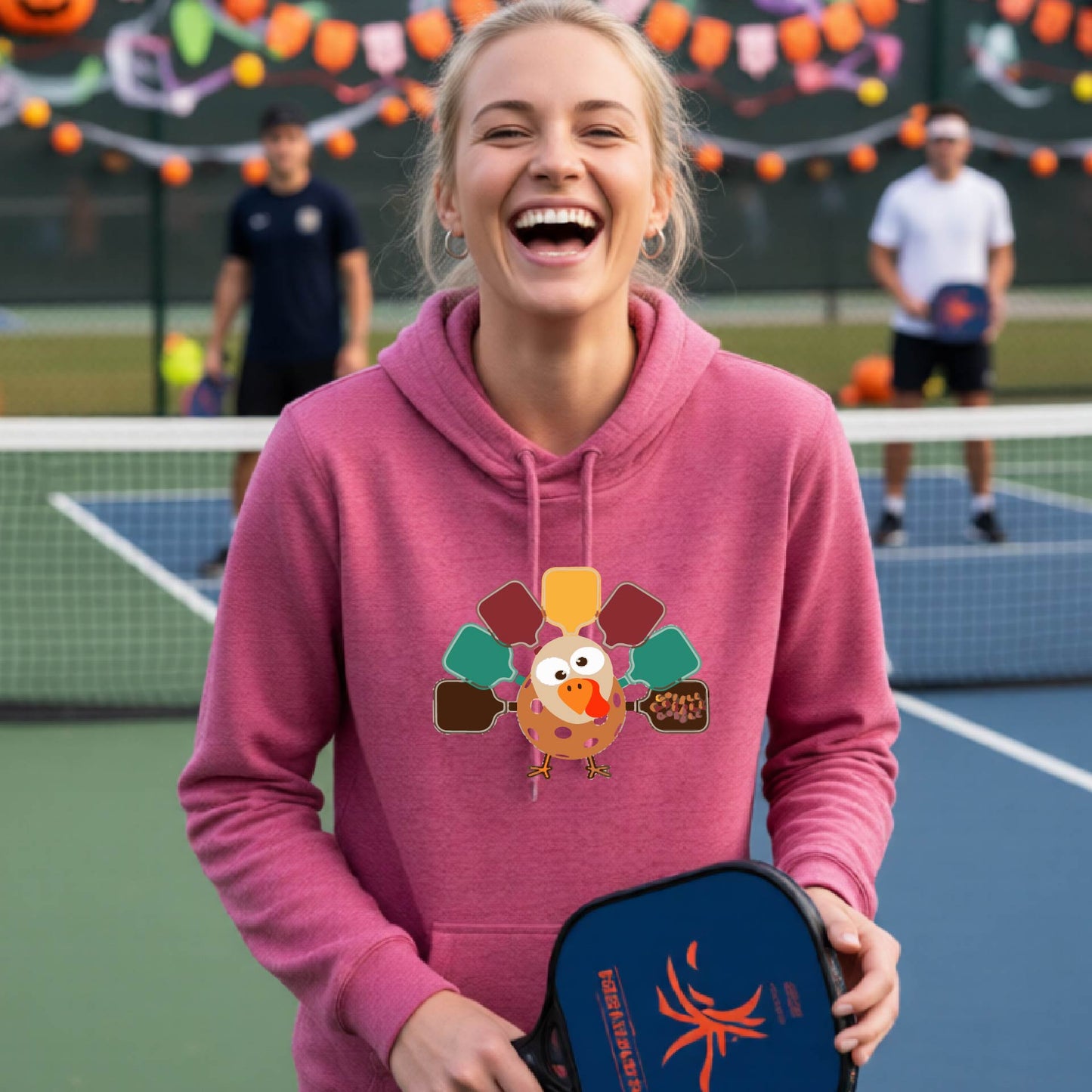A woman smiling in a pickleball court wearing a  pink hoodie with a colorful pickleball turkey graphic made of pickleball paddles on the 