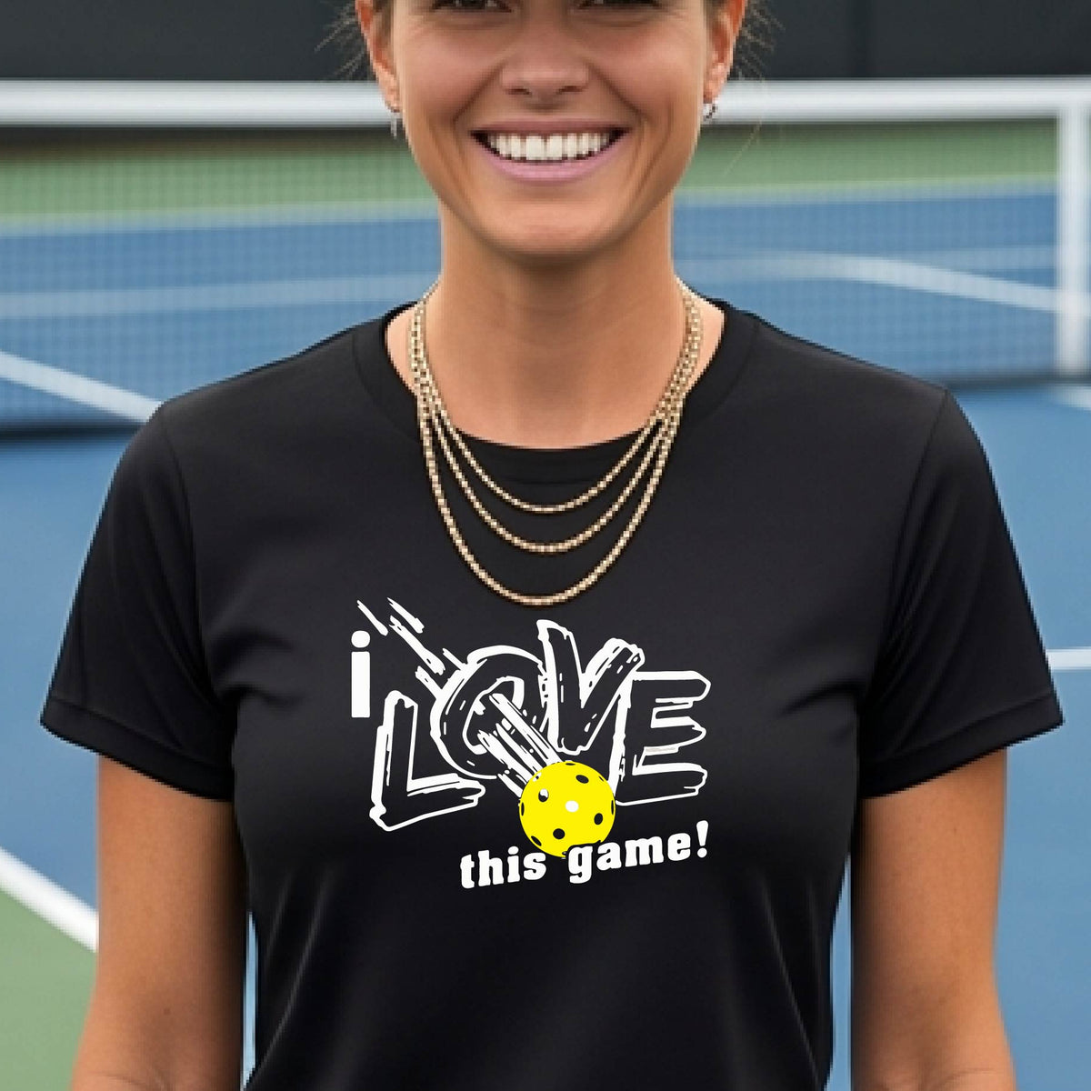 A woman on a pickleball court wearing a black short-sleeve t-shirt with a graphic that reads 'i LOVE this game!' in white, with a yellow pikleball illustration.