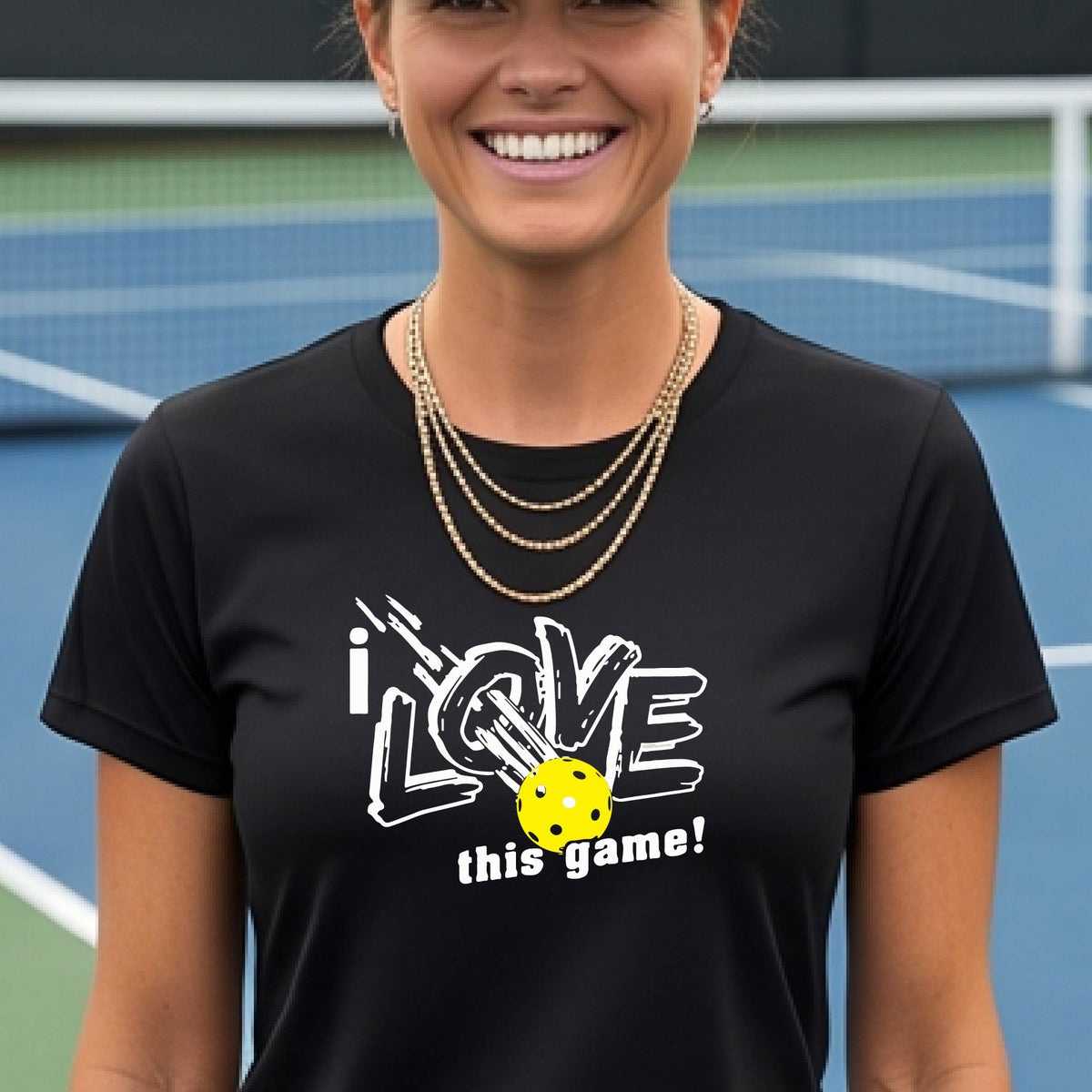 A woman on a pickleball court wearing a black short-sleeve t-shirt with a graphic that reads 'i LOVE this game!' in white, with a yellow pickleball illustration.