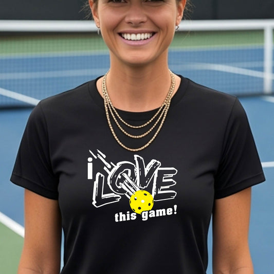 A woman on a pickleball court wearing a black short-sleeve t-shirt with a graphic that reads 'i LOVE this game!' in white, with a yellow pickleball illustration.