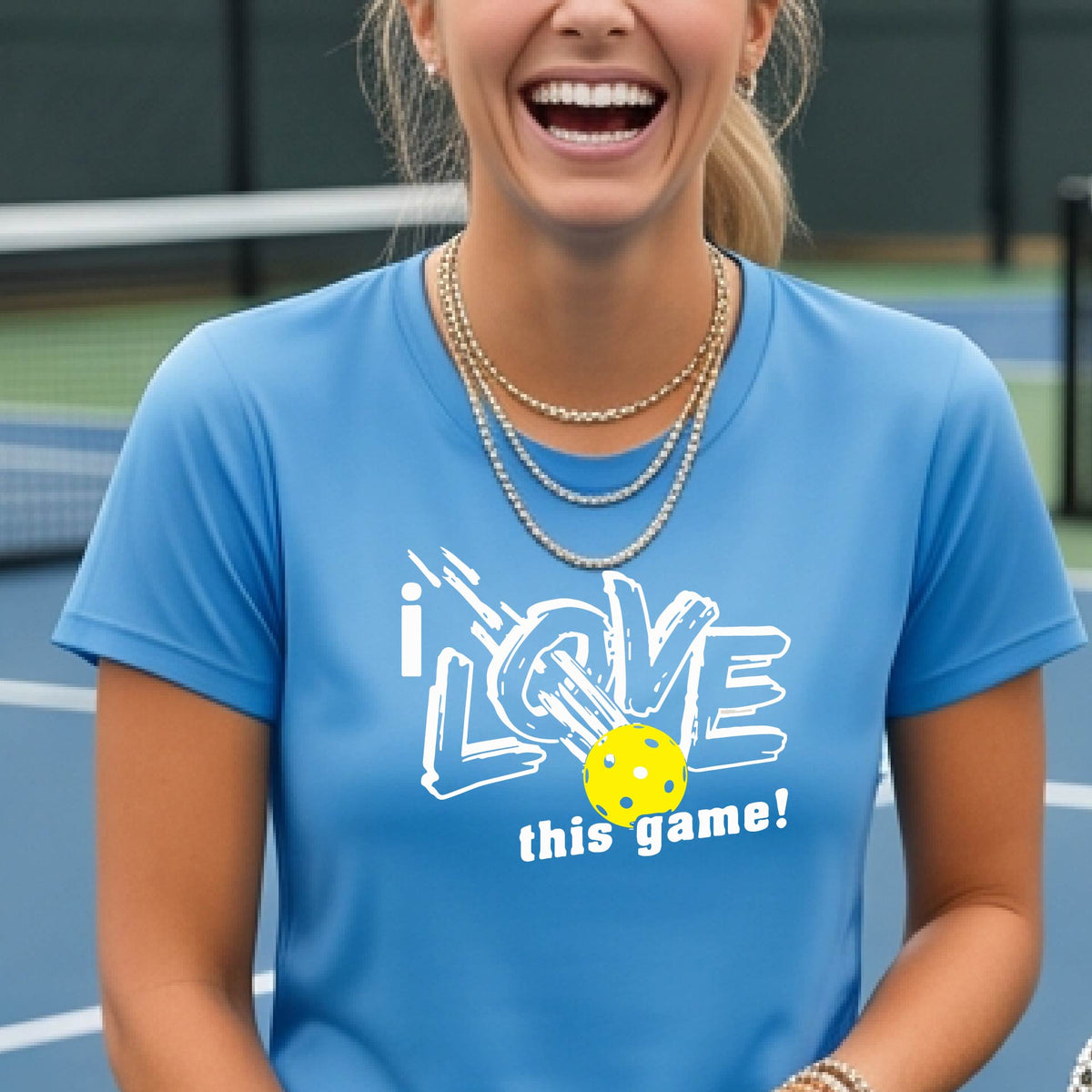 A woman on a pickleball court wearing a light blue short-sleeve t-shirt with a graphic that reads 'i LOVE this game!' in white, with a yellow pickleball illustration.