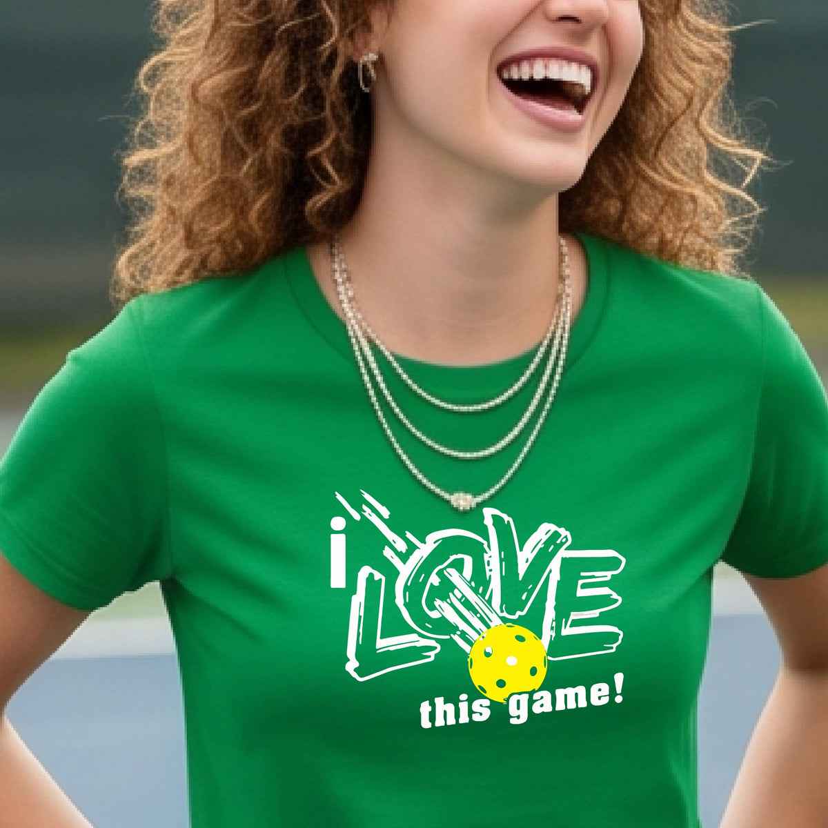 A woman on a pickleball court wearing a green short-sleeve t-shirt with a graphic that reads 'i LOVE this game!' in white, with a yellow pikleball illustration.