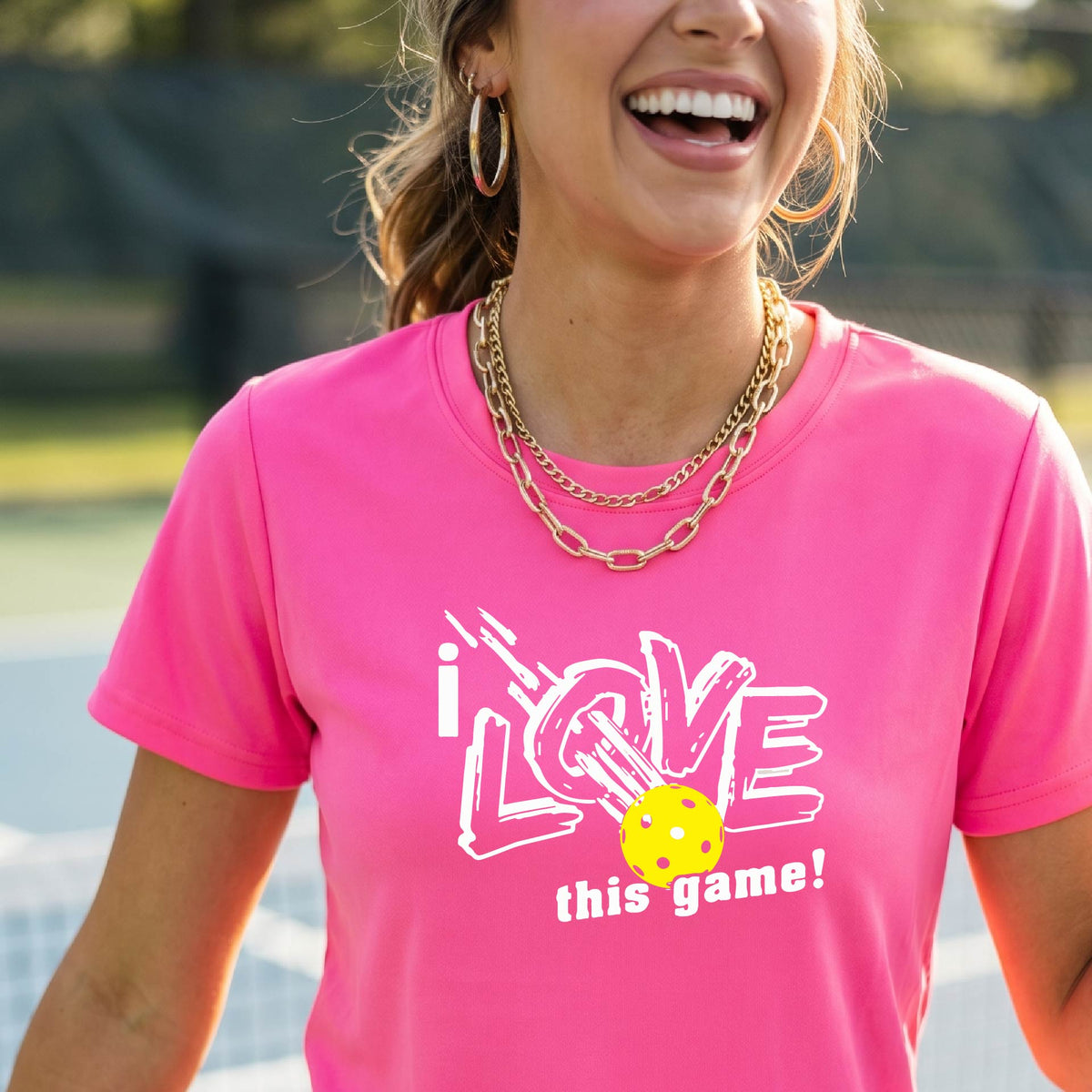 A woman on a pickleball court wearing a pink short-sleeve t-shirt with a graphic that reads 'i LOVE this game!' in white, with a yellow pikleball illustration.