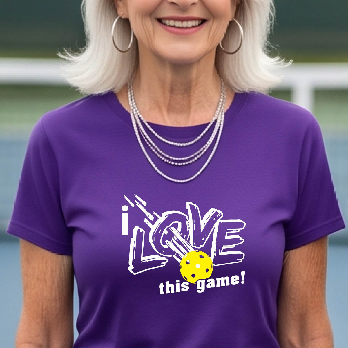 A woman on a pickleball court wearing a purple short-sleeve t-shirt with a graphic that reads 'i LOVE this game!' in white, with a yellow pikleball illustration.