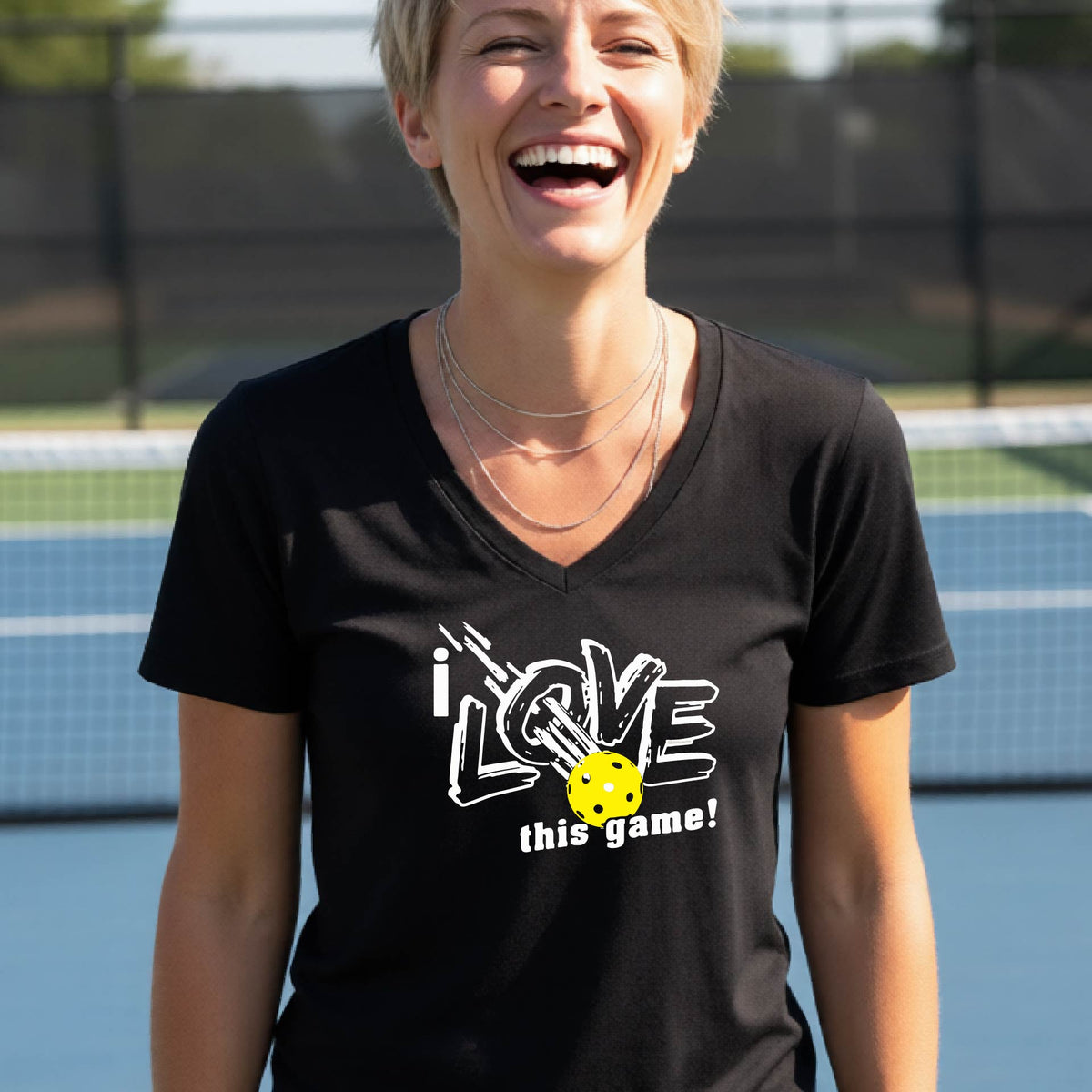 A smiling woman on a pickelball court wearing a black V-neck t-shirt with a graphic that reads 'i LOVE this game!' with white and yellow pickleball.