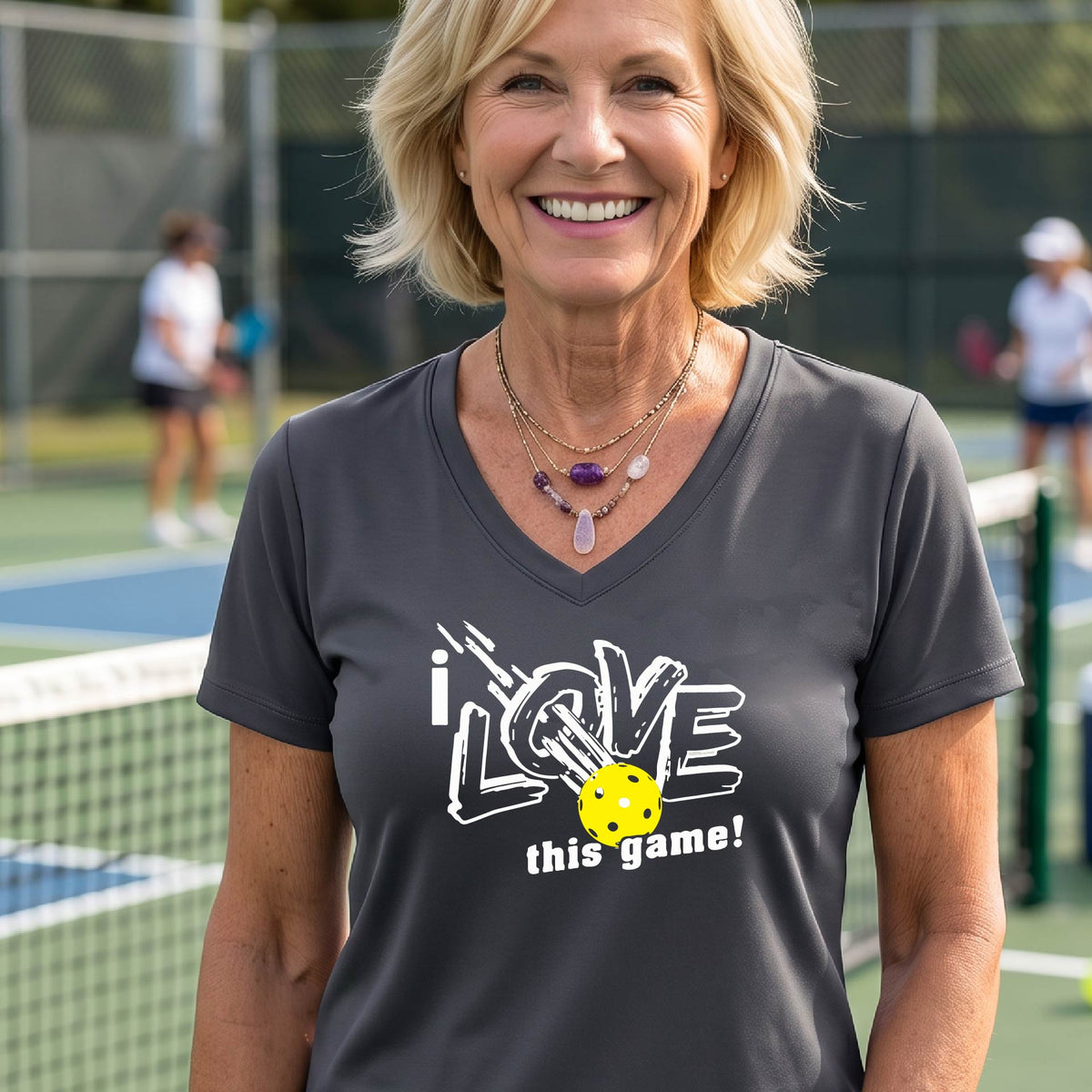 A smiling woman on a pickelball court wearing a gray V-neck t-shirt with a graphic that reads 'i LOVE this game!' with white and yellow pickleball.