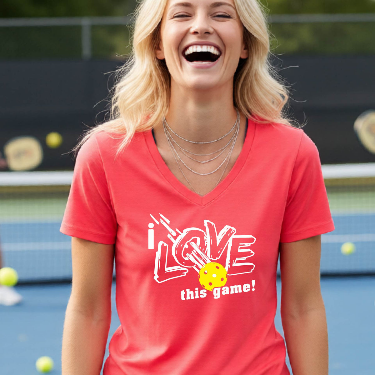 A smiling woman on a pickelball court wearing a coral pink V-neck t-shirt with a graphic that reads 'i LOVE this game!' with white and yellow pickleball.
