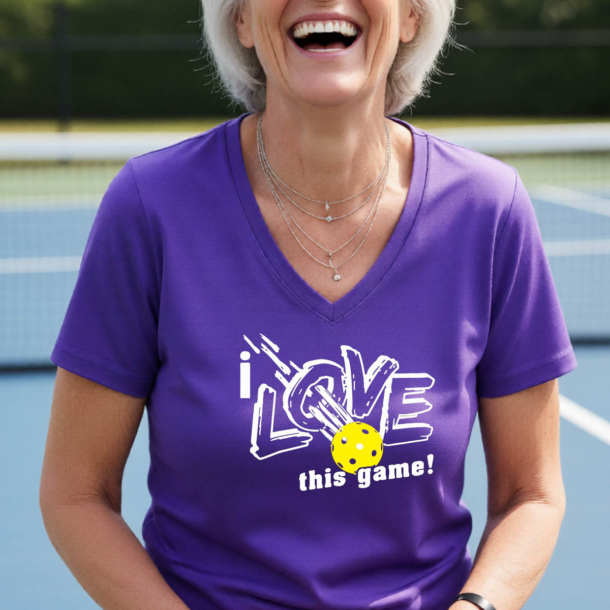 A smiling woman on a pickelball court wearing a purple V-neck t-shirt with a graphic that reads 'i LOVE this game!' with white and yellow pickleball.