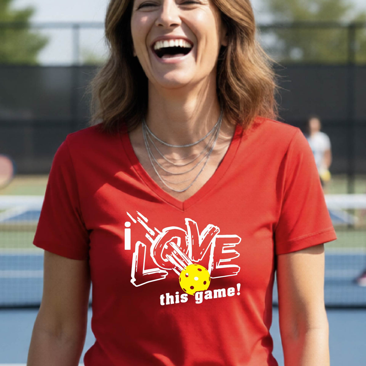 A smiling woman on a pickelball court wearing a red V-neck t-shirt with a graphic that reads 'i LOVE this game!' with white and yellow pickleball.