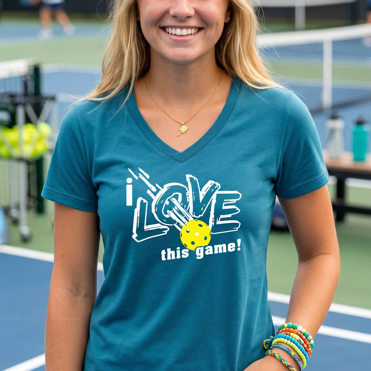 A smiling woman on a pickelball court wearing a teal V-neck t-shirt with a graphic that reads 'i LOVE this game!' with white and yellow pickleball.