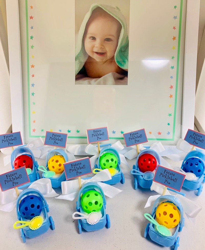 Pickleball Baby Shower Favors - Set of Eight Tiny Carriages Baby with Micro Pickleballs  For all the future pickleball pros, these baby shower favors are a must have. The stroller measures only 2.5" high and long and 1.5" wide and the pickleball baby is accented with a charming baby rattle and beautiful white bow.  These adorable favors are sure to delight all your pickleball friends and are the prefect addition to your celebration. Show your love of pickleball and spice up the 